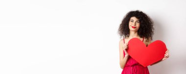 Genç romantik kadın sevgililer gününde aşkı düşlüyor, ruh eşi arıyor, büyük kırmızı bir kalp kesiği tutuyor ve soldan dalgın dalgın bakıyor, beyaz bir arka plan..