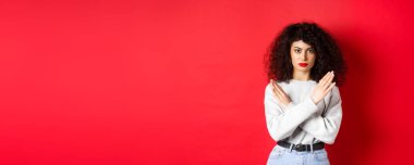 Kıvırcık saçlı, ciddi bir beyaz kadın hayır diyor. Kırmızı arka planda hoşnutsuz duran, kötü bir şeyi engellemek için çapraz jest yapıyor..