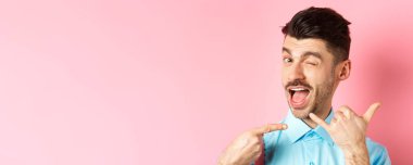 Küstah ve komik adam telefon işareti yapıyor, onu aramak istiyor ve göz kırpıyor, numarasını söylüyor, pembe arka planda duruyor..