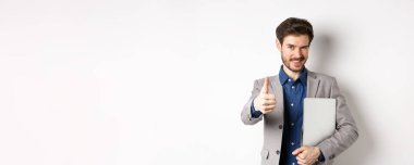 Başarılı bir iş adamı şık bir takım elbiseyle dizüstü bilgisayar taşır ve kameraya kendinden emin bakar, onaylı, beyaz arka planda başparmak kaldırır..