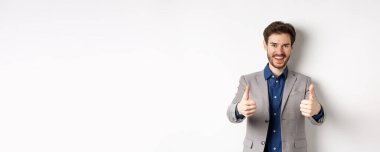 Takım elbiseli neşeli ve pozitif genç adam iyi bir şey gibi başparmaklarını kaldırıyor ve gülümsüyor, iyi bir iş çıkarıyor, beyaz arka planda mutlu duruyor..