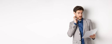 Kafası karışmış, takım elbiseli, sakallı, cahil, evrakları anlamayan, elinde kararsız yüzlü, beyaz arka planlı bir kağıt tutan bir ofis çalışanı..