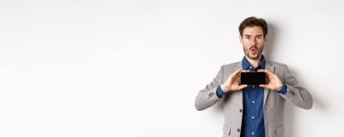 E-ticaret ve online alışveriş konsepti. Heyecanlı takım elbiseli genç adam boş akıllı telefon ekranı gösteriyor ve hayret verici, beyaz arka plan diyor..