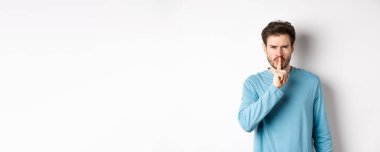 Sinirli adam kameraya şşşş yapıyor ve sessiz olmasını söylüyor, üzgün, beyaz arka plan üzerinde duruyor dudaklarında parmak, tabu jesti.