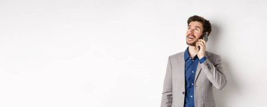 Tasasız genç adam cep telefonuyla konuşuyor ve gülümsüyor, rüya gibi bir yüzle yukarı bakıyor, beyaz arka planda takım elbise içinde duruyor..