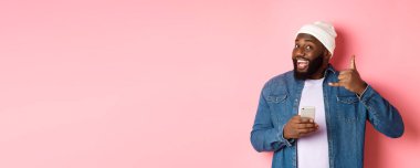 Mutlu siyah adam bana işaret veriyor, telefon ediyor, gülümsüyor, akıllı telefon tutuyor, bereyle ve kot gömlekle pembe arka planda dikiliyor..