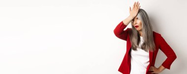 Kırmızı ceketli ve facepalmlı sinirli Asyalı ofis hanımı, beyaz arka planda rahatsız ve tedirgin duruyor..