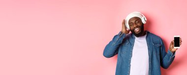 Kaygısız yakışıklı siyah adam kulaklıkla müzik dinliyor, sesin keyfini çıkarıyor ve akıllı telefon ekranı gösteriyor, pembe arka planda yüzünde mutlu bir gülümsemeyle duruyor..