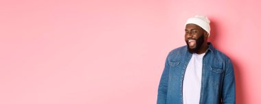 Arsız Afro-Amerikalı adam kameraya göz kırpıyor ve gülümsüyor, teklifle dalga geçiyor, pembe arka plana karşı hippi kıyafetleri içinde duruyor..