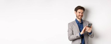 Gülümseyen başarılı iş adamı beyaz arka planda akıllı telefon kullanıyor, takım elbise içinde duruyor..