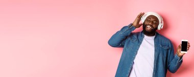 Mutlu Afro-Amerikalı hipster adam kulaklıkla müzik dinliyor, telefon ekranı uygulamasını gösteriyor ve birlikte şarkı söylüyor, pembe arka planda duruyor..