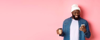 Tarz sahibi siyahi bir hippinin kahve içerken, telefonla mesaj okurken, gülümserken, pembe arka planda dururken görüntüsü..