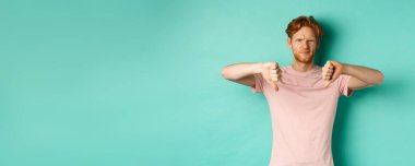 Hayal kırıklığına uğramış kızıl saçlı, baş parmağını kaldırmış, kaşlarını çatmış, şüpheci, hoşnutsuz ve hoşnutsuz görünen, turkuaz arka planda duran bir adam..