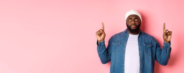 Endişeli ve tereddütlü siyah adam, bereli parmaklarını havaya kaldırarak, şüpheli yüzüyle, pembe arka planda dikiliyor..