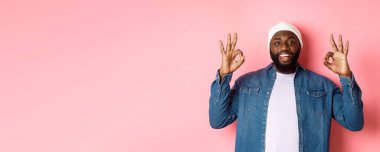 Hipster bereli tatmin olmuş Afro-Amerikan bir adam, onaylama işaretleri gösteriyor, iyi bir teklifi övüyor ve evet diyor, mutlu gülümsüyor, pembe arka planda duruyor..