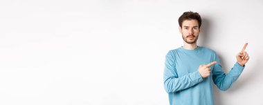 Beyaz arka planda sıradan kıyafetler giymiş, logoyu işaret eden ve kameraya bakan ciddi bir beyaz adamın portresi..