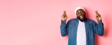 İyimser Afro-Amerikalı bir adam dilek diliyor, parmaklarını çapraz tutuyor ve gülümsüyor, pembe arka planda duruyor..