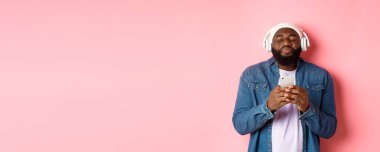 Harika müziğin keyfini çıkaran, kulaklıkla şarkı dinleyen, akıllı telefon tutan, kendinden geçmiş, pembe arka planda duran siyahi bir adam..