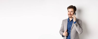 Başarılı iş adamı takım elbisesini onarıyor ve cep telefonuyla konuşuyor. Kameraya gülümsüyor, beyaz arka planda duruyor..