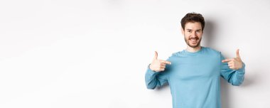 Yakışıklı sakallı adam, parmaklarını merkeze doğrultuyor, kendi reklamını yapıyor ve kendinden emin görünüyor, beyaz arka planda duruyor, kameraya gülümsüyor..