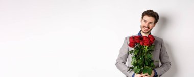 Yakışıklı, güler yüzlü bir adam romantik bir randevuya çıkıyor, takım elbise giyiyor, elinde gül buketi tutuyor ve sola bakıyor, sevgililer gününde beyaz arka planda hediyesiyle dikiliyor..