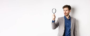 Takım elbiseli ciddi bir adam büyüteçle ipucu arıyor, araştırıyor, beyaz arka planda duruyor..