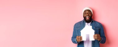 Emlak konsepti. Mutlu gülümseyen Afro-Amerikan adam ev modelini tutuyor, kameraya heyecanlı bakıyor, evini arıyor, pembe arka planda duruyor..