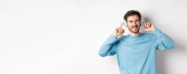 Çekici, sakallı, gülümseyen, müzik dinlerken kulağına dokunan, beyaz arka planda duran modern bir adam..