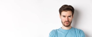 Suratsız, somurtkan ve gücenmiş suratlar yapan, beyaz arka planda üzgün duran huysuz genç adamın yakın çekimi..