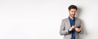 Yakışıklı sakallı erkek model cep telefonu kullanıyor, kameraya gülümsüyor, beyaz arkaplan.