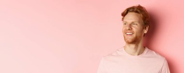 Sakallı, kızıl saçlı, soluna bakıp gülümseyen pembe arka planda kendine güvenen yakışıklı bir genç..