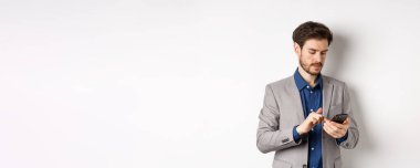 Şık takım elbiseli yakışıklı iş adamı akıllı telefonda konuşuyor, cep telefonu kullanıyor, beyaz arka planda duruyor..