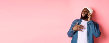 Mutlu Afro-Amerikalı adam cep telefonuyla konuşuyor, gülüyor ve gülümsüyor, pembe arka plan üzerinde bere ve kot gömlek ile duruyor..