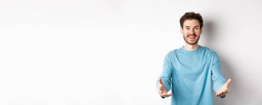 Gündelik giysiler içindeki çekici beyaz adam bir şeyi tutmak için ellerini uzatıyor, gülümsüyor ve alıyor, beyaz arka planda duruyor..