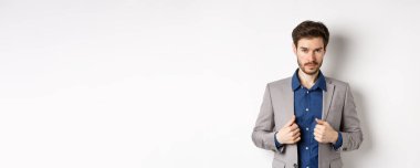 Sakal düzeltme kıyafeti giymiş, kameraya küstah bakışlı, hazır ve kararlı, beyaz arka planda duran kendine güvenen genç bir adam..