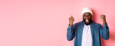 Mutlu siyahi adamın piyangoyu kazanması, sevinmesi ve yumruk tokuşturması, sevinçle evet diye bağırması, pembe arka planda durması..