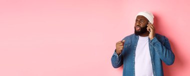 Kızgın Afro-Amerikalı adam telefonda konuşuyor ve birini yumruğuyla tehdit ediyor, rahatsız bir şekilde bakıyor, pembe arka planda duruyor..