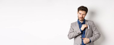 Kendine güvenen ve başarılı iş adamı bir kenara çekilirken kredi kartını takım elbisenin cebine koyup beyaz arka planda duruyor..