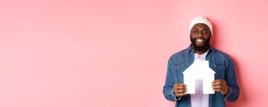 Emlak konsepti. Mutlu gülümseyen Afro-Amerikalı adam ev modelini tutuyor, daire alıyor, pembe arka planda duruyor..