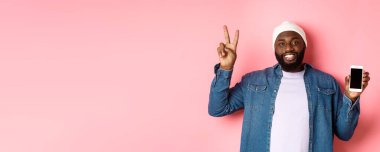 Çevrimiçi alışveriş ve teknoloji kavramı. Bereli ve kot gömlekli mutlu siyah adam pembe arka planda duran mobil ekran ve barış işareti gösteriyor..