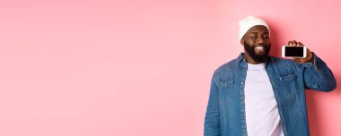 Çevrimiçi alışveriş ve teknoloji kavramı. Mutlu genç Afro-Amerikalı adam gülümsüyor, akıllı telefon ekranını yatay olarak gösteriyor, tatmin olmuş yüz ifadesi, pembe arka plan.