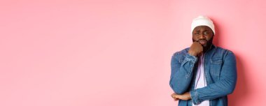 Genç Afro-Amerikalı adam kameraya acıyarak ve endişeyle bakıyor, kaşlarını çatmış ve tedirgin, pembe arka planda duruyor..