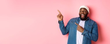 Şaşırmış siyahi adam harika bir reklam sunuyor, gülümsüyor ve parmaklarını sol üst köşedeki pankarta gösteriyor, pembe arka plan..
