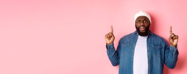 Hayal kırıklığına uğramış ve üzgün siyah adam kaşlarını çatmış, parmaklarını kaldırmış ve kameraya bakıyor rahatsız, şikayet ediyor, pembe arka planda duruyor..