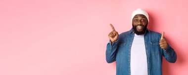 Mutlu Afro-Amerikalı adam baş parmağını yukarı kaldırıyor ve sol üst köşedeki fotokopi alanını işaret ediyor, iyi bir anlaşmaya övgüler yağdırıyor, pembe arka planda duruyor..