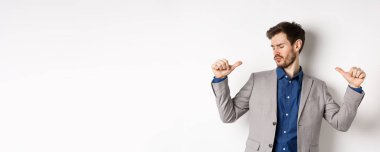 Takım elbiseli yakışıklı ve kendine güvenen bir adam kendini gösterip havalı ve küstah hissediyor, iyi görünümünü tanıtıyor, beyaz arka planda duruyor..