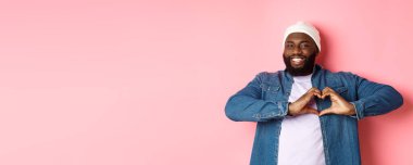 Mutlu Afro-Amerikalı adam kalp işareti gösteriyor, seni seviyorum, jest yapıyorsun, kameraya gülümsüyorsun, pembe arka plan.