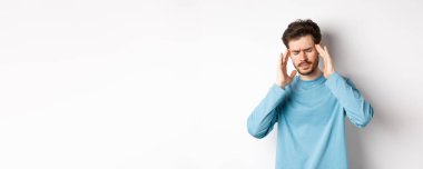 Genç adam baş ağrısı çekiyor ya da akşamdan kalma, baş şakaklarına dokunuyor ve kaşlarını çatıyor, migrenden midesi bulanıyor, beyaz arka planda duruyor. .