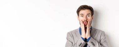 Şaşkın takım elbiseli işadamı, inkar edilmiş, dudakları büzüşmüş, yüzüne dokunmuş, kameraya bakıyor, beyaz arka planda duruyor..