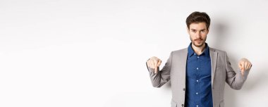 Bana bak. Takım elbiseli yakışıklı sakallı adam, parmaklarını aşağı gösteriyor, kameraya bakıyor, afişi gösteriyor, beyaz arka planda duruyor..